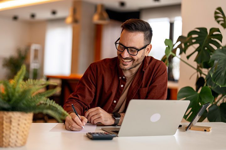 A person wearing glasses and a red shirt is smiling while writing at a desk with a laptop, tablet, and calculator. A plant is nearby.