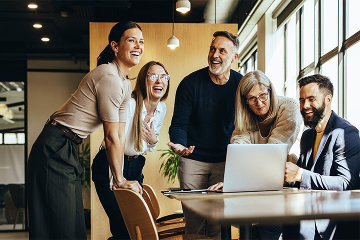 A group of five adults in a modern office laugh and discuss something around a laptop on a table. Large windows let in natural light.