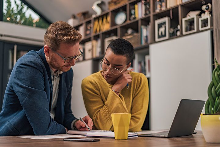 Two people work together at a table, one writing in a notebook. A laptop, yellow mug, and smartphone are on the table. Shelves with books and decor are in the background.