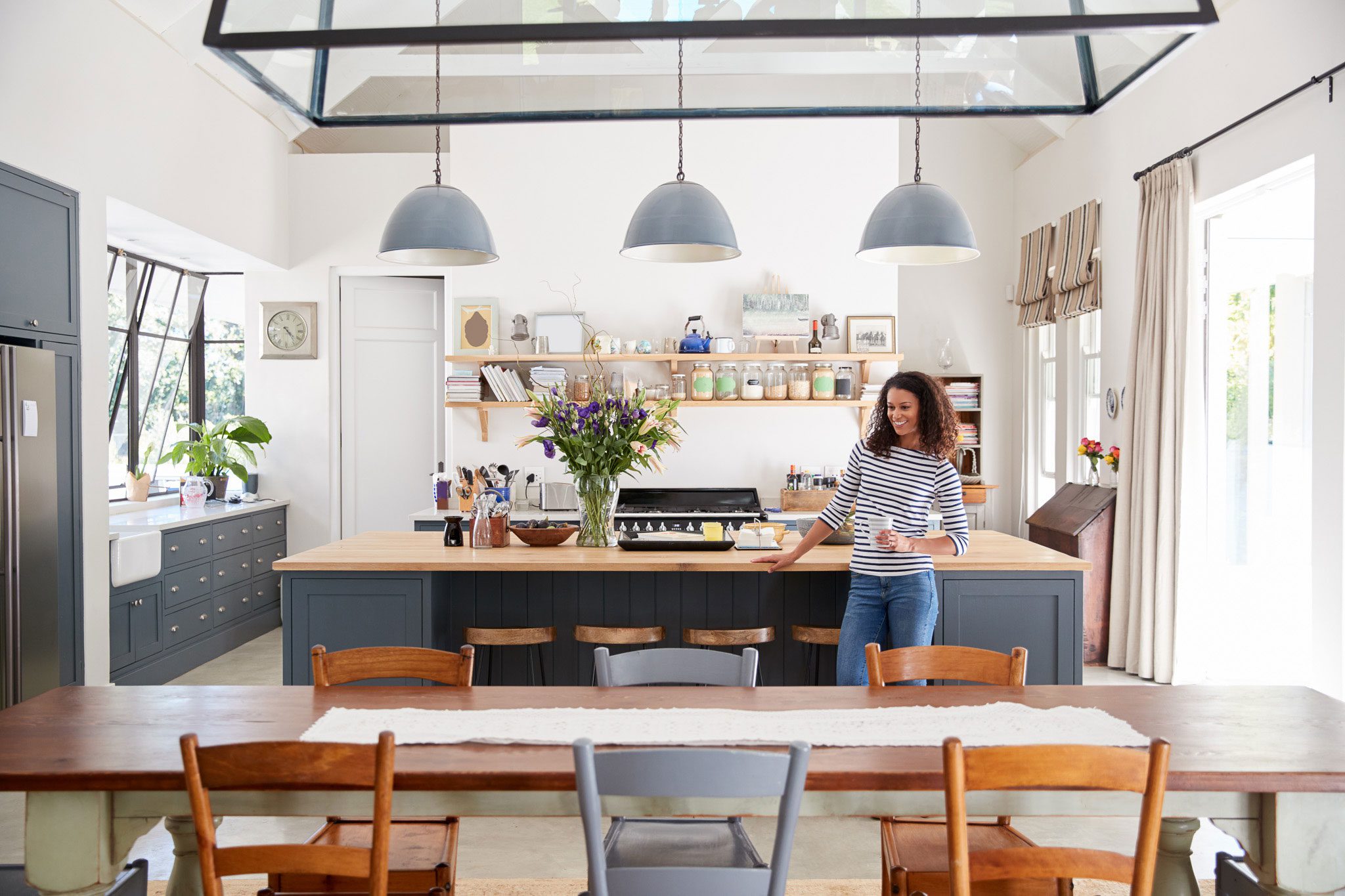 A woman stands in a bright, modern kitchen holding a mug, near an island with flowers and shelves filled with jars and kitchenware.