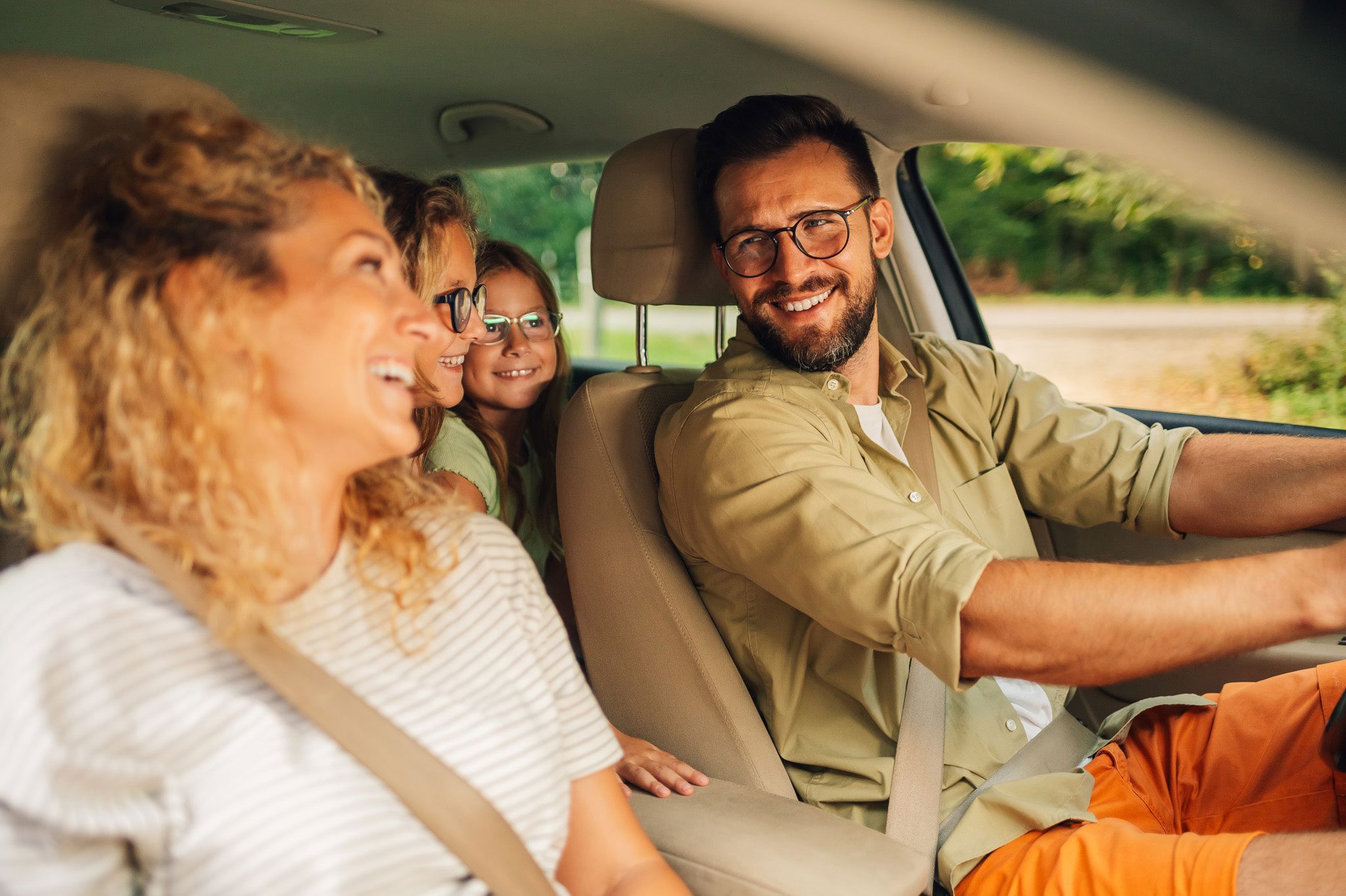 A man drives a car with a woman in the front passenger seat and two children in the back, all smiling and wearing seatbelts.