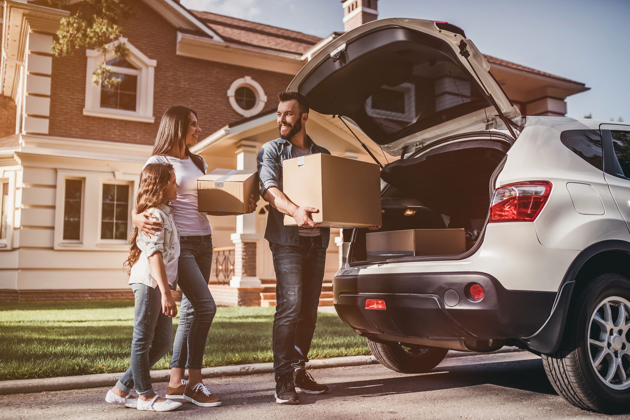 A family stands by a car with its trunk open, holding cardboard boxes in front of a suburban house, suggesting moving or unpacking.
